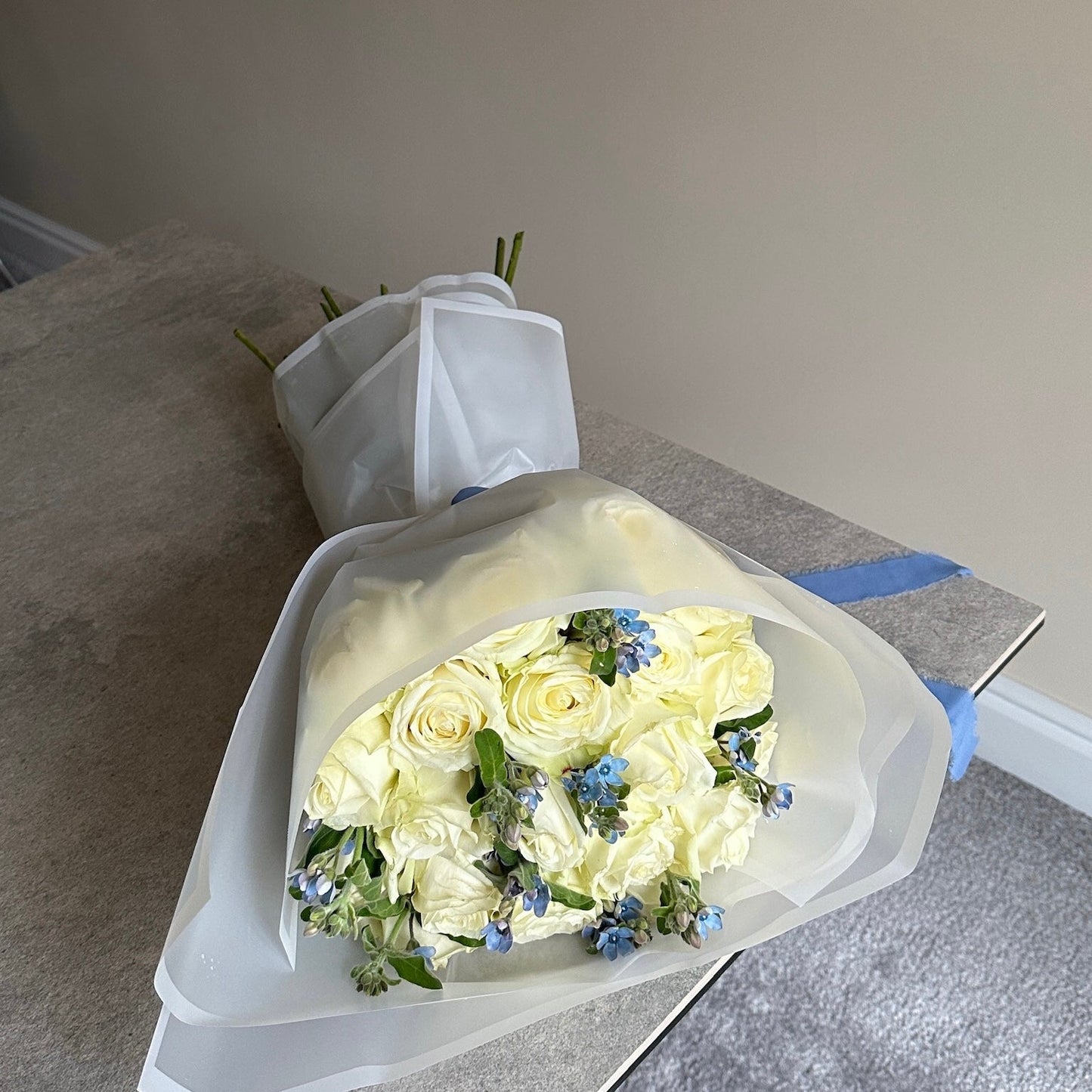 Bouquet of white roses with a few blue flowers in white cloud wrap on a gray surface.