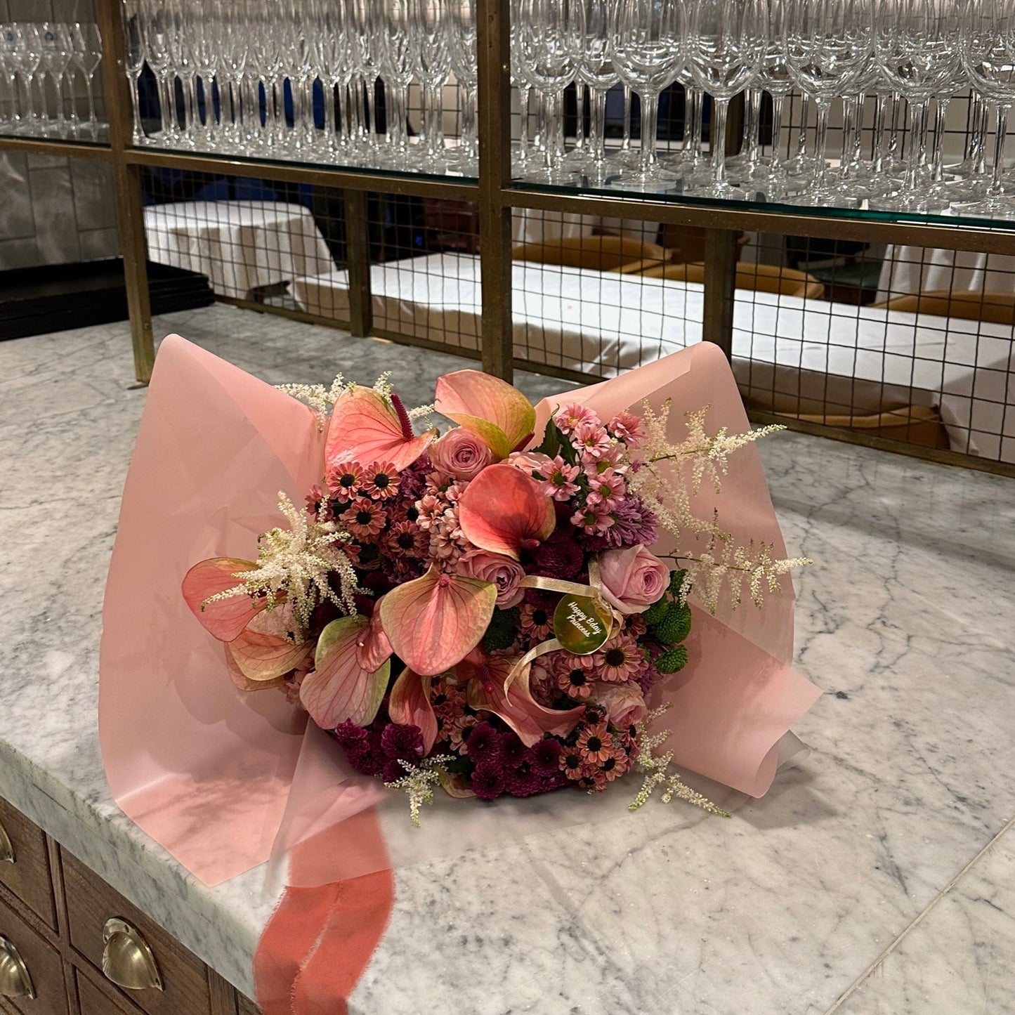 Bouquet of flowers wrapped in pink paper on a marble countertop with glass shelves in the background.