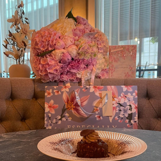 Floral arrangement with hydrangeas and peonies in velour wrapping and a piece of cake on a table with a decorative box in the background.