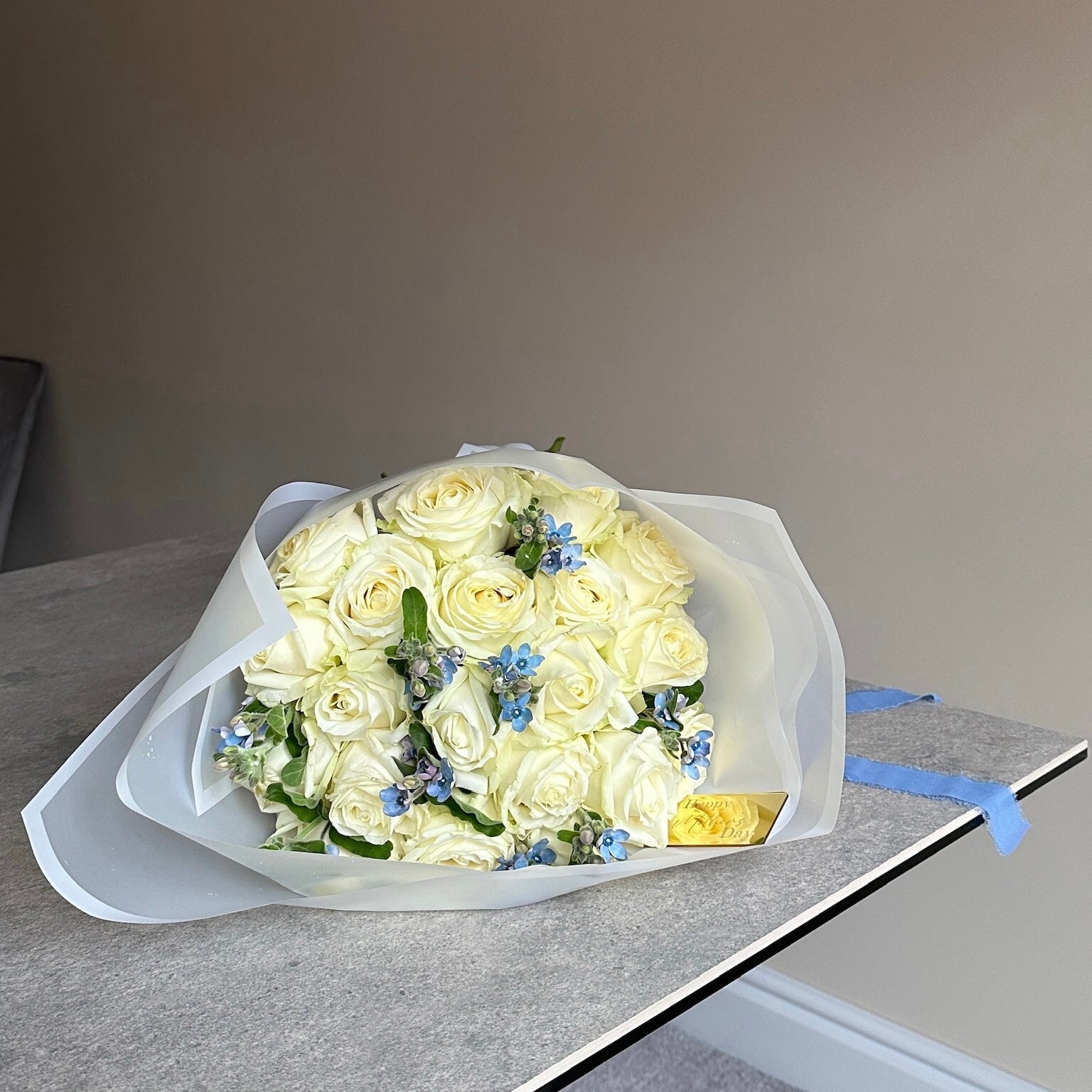 Bouquet of white roses with a few blue flowers in white cloud wrap on a gray surface.