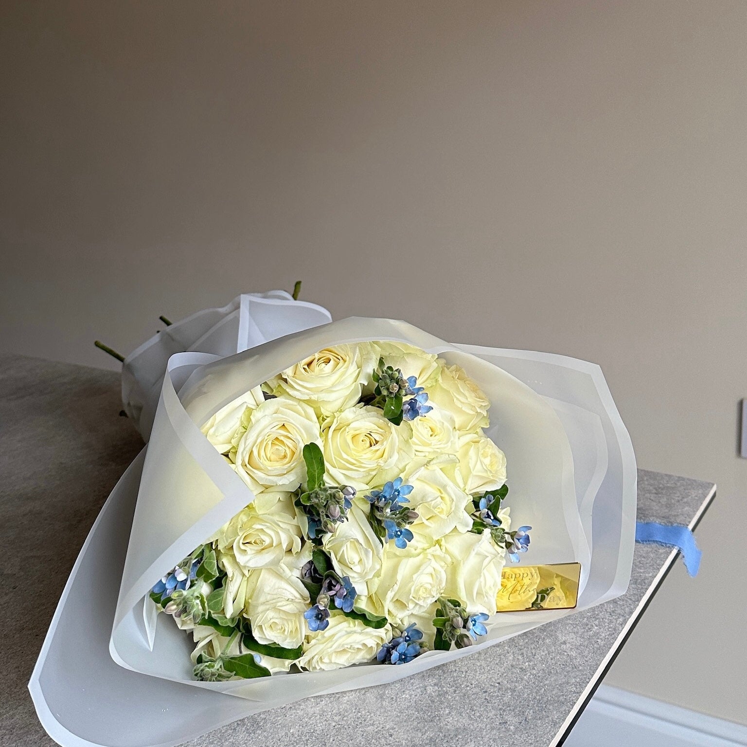 Bouquet of white roses with a few blue flowers in white cloud wrap on a gray surface.