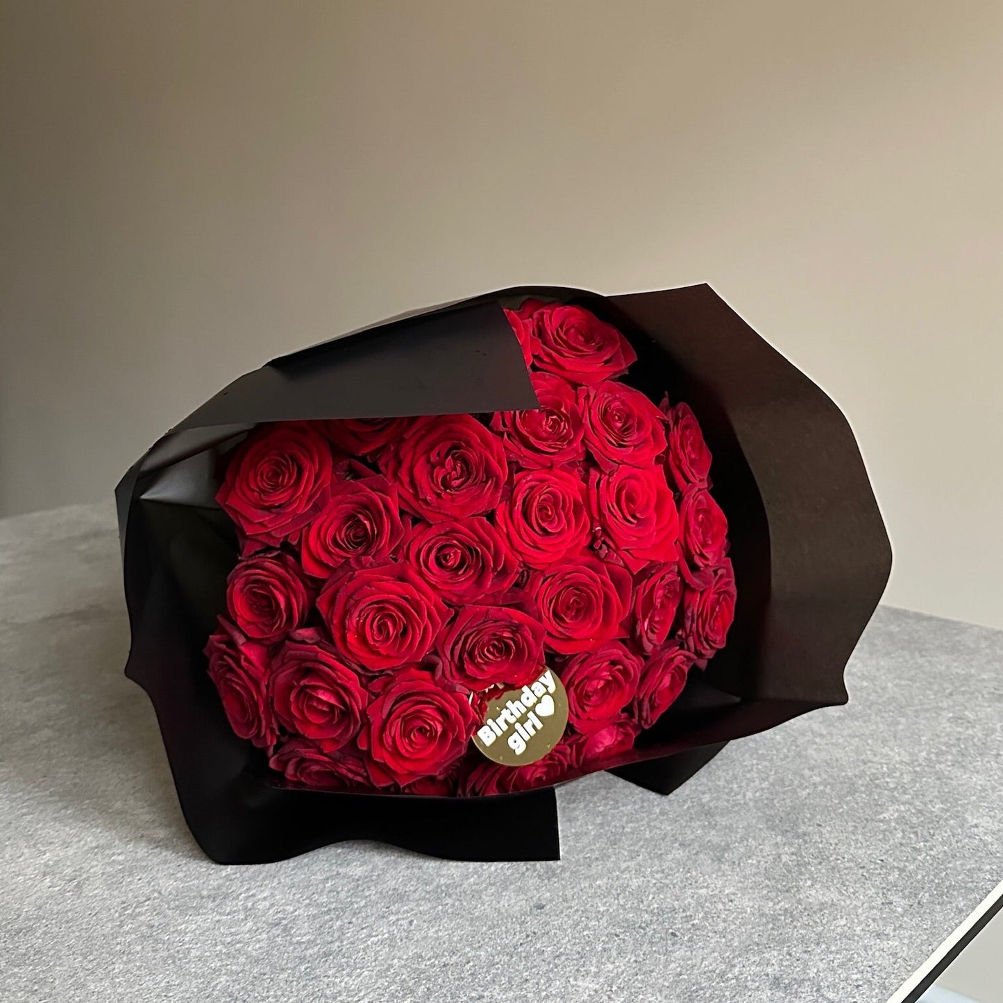 Bouquet of red roses wrapped in black paper, with a personalised message on a gold tag, cloud black wrapping on a light gray surface with a neutral background