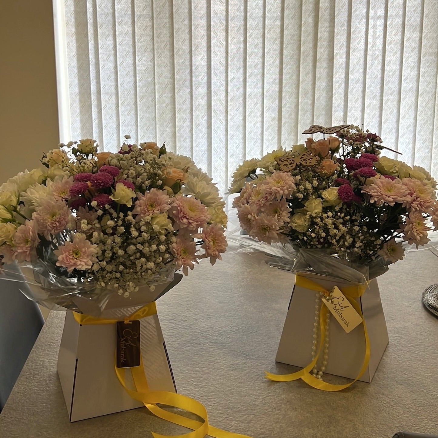 Two bouquets of flowers in a box with yellow ribbons in front of a window with blinds.