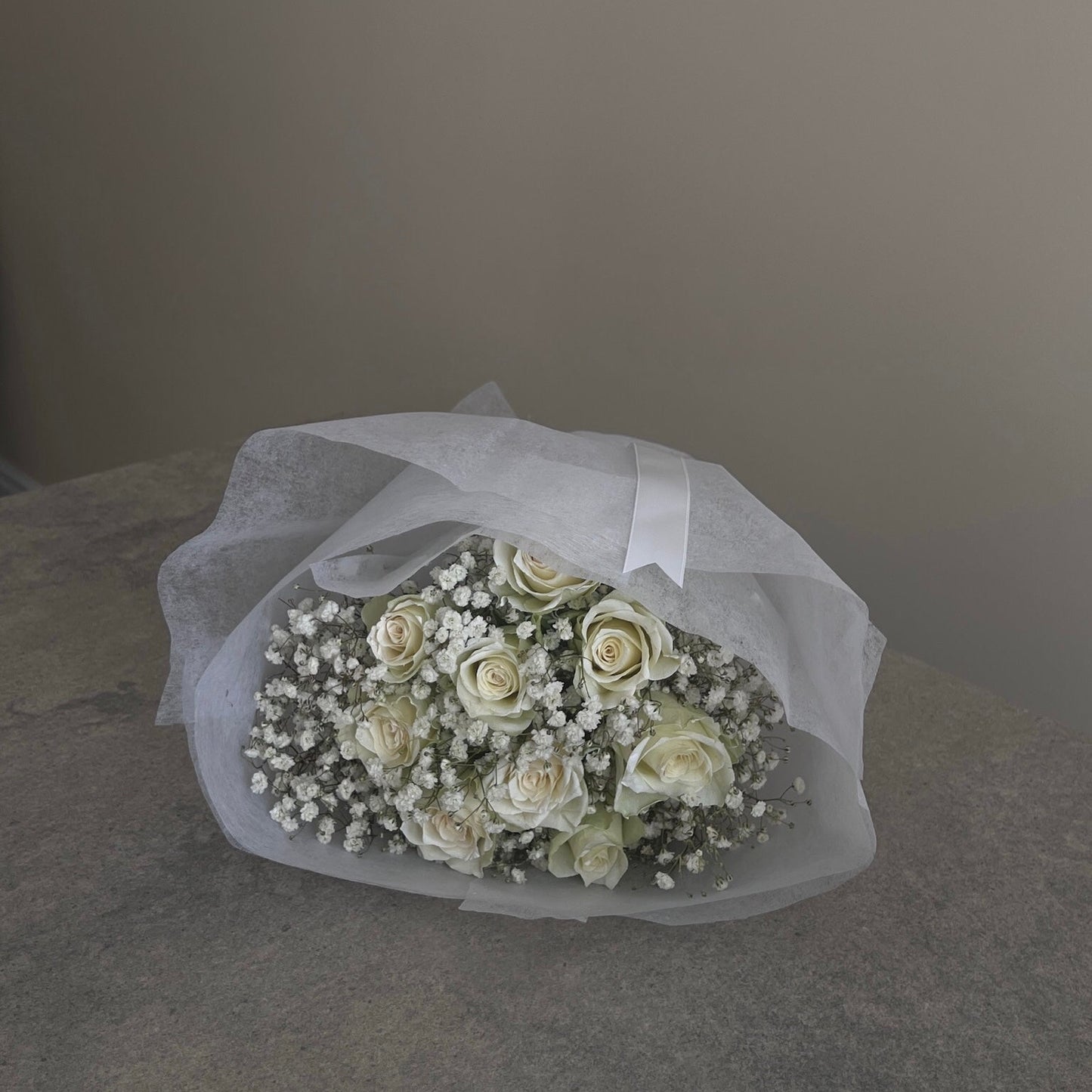 Bouquet of white roses and baby's breath wrapped in tissue paper on a gray surface.